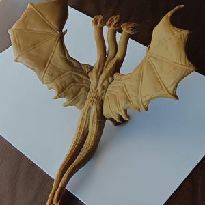 Può includere: Una statuetta di drago a tre teste color marrone chiaro, con grandi ali dettagliate. Il drago ha una lunga coda ed è posizionato su una superficie bianca. La statuetta è probabilmente realizzata in resina o materiale simile.