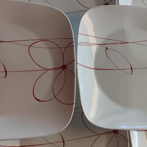 May include: White square plates with a red floral design. The plates have rounded corners and a glossy finish. The red design features a stylized flower with curved lines and a central dot. A set of plates for dining.