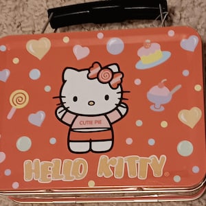 May include: A red Hello Kitty lunchbox with a black handle. The lunchbox features a Hello Kitty graphic with the words "Cutie Pie" on her shirt. The design includes hearts, lollipops, and desserts.