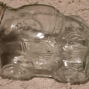 Op de afbeelding: Een doorzichtige glazen pot in de vorm van een oldtimer, met gegoten details van wielen, ramen en een motorkap. De pot heeft een brede opening aan de bovenkant, die is afgesloten met een ronde, lichtbruine kurken stop.