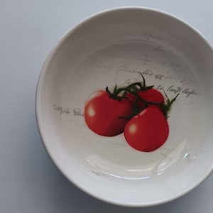 May include: White ceramic bowl with a red tomato design. The bowl features three ripe tomatoes with green stems. The design is set against a backdrop of faded script. A kitchenware item.