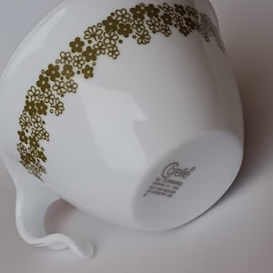 Könnte beinhalten: Weiße Corelle-Tasse mit gebogenem Griff, verziert mit einem Muster aus kleinen, olivgrünen Blumen. Die Tasse hat eine glänzende Oberfläche und das Corelle by Corning-Logo ist unten sichtbar.