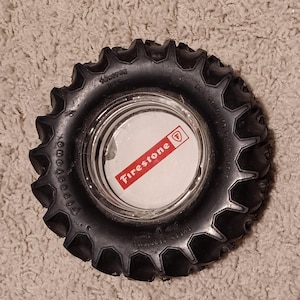 May include: Black rubber ashtray in the shape of a tractor tire. The center features a white circle with the red Firestone logo. The tire has a textured surface and is designed with deep treads.
