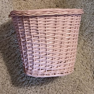 Può includere: Un cestino intrecciato rosa chiaro con una forma leggermente svasata. Il cestino è realizzato con strisce intrecciate, creando una superficie strutturata. Il bordo superiore è rifinito con un bordo arrotolato. Il cestino viene probabilmente utilizzato per riporre oggetti o per decorare.