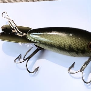 May include: A vintage fishing lure with a dark green and black body, featuring two treble hooks and a silver spinner. The lure is next to a U.S. quarter for size comparison. The lure has a detailed scale pattern and a gold-colored eye.
