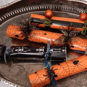 May include: A selection of Halloween crackers on a silver tray. The crackers are orange with black spiderwebs and spiders, and black with white skeleton designs. Some are decorated with orange baubles and black spider accents.