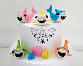 May include: A two-tiered white cake decorated with fondant shark and fish figurines. The top tier features three sharks in pink, yellow, and blue. The bottom tier has two sharks in green and orange, along with coral and seaweed decorations. The cake is topped with the text "Fancy Sugar on Top".