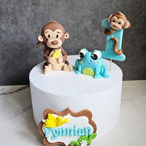 May include: A cake topper featuring a brown monkey holding bananas, a blue frog, and a monkey on a blue number one. A brown and white plaque with the name "Rynnian" in blue letters and floral accents is also included.