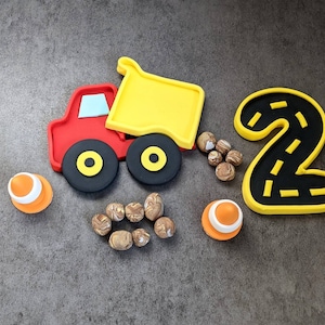 May include: A yellow toy dump truck with a red cab and black wheels is carrying brown rocks. A yellow number two with a black road design is next to the truck. Two orange traffic cones are on either side of the truck.