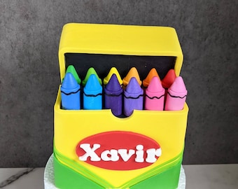 May include: A yellow and green cake decorated to look like a box of crayons. The box is open and has a row of colorful crayons inside. The name "Xavier" is written on the front of the box.