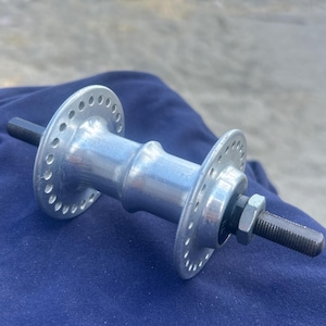 Op de afbeelding: Een zilverkleurige metalen fietswielnaaf met twee ronde flenzen met meerdere spaakgaten, een centraal cilindrisch lichaam en een zwarte metalen as met schroefdraad die aan beide uiteinden uitsteekt met een zeskantmoer.