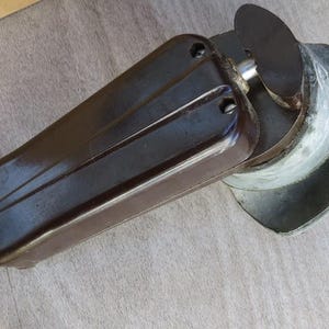 Könnte beinhalten: Ein dunkelbrauner, rechteckiger Griff aus geformtem Kunststoff, der an einem metallischen, halbkreisförmigen Bauteil befestigt ist. Der Griff hat zwei sichtbare Schraubenlöcher und eine glatte, glänzende Oberfläche, während das Metallteil abgenutzt wirkt.
