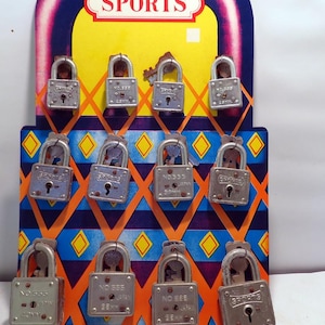 Puede incluir: Una tarjeta de exhibición comercial con la palabra SPORTS en la parte superior, que contiene doce candados de metal plateado de varios tamaños. Cada candado tiene las inscripciones SPORTS, NO.555 y JAPAN, con una llave en el arco.