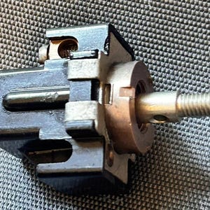 以下が含まれることがあります： 黒い電気ポテンショメータ部品。長方形のハウジング、中央の金属製ネジ付きシャフト、露出した銅色の内部接点が特徴です。このデバイスには、銀色の取り付けカラーと小さな側面スロットが含まれています。