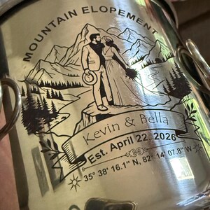 May include: A silver-toned metal mug featuring a black engraved illustration of a couple standing before mountains. The text reads MOUNTAIN ELOPEMENT, Kevin & Bella, Est. April 22, 2026, and the coordinates 35° 38' 16.1" N, 82° 14' 07.8" W.