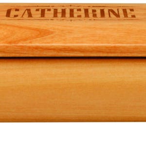 May include: A light-coloured wooden jewellery box with a rectangular shape and rounded edges. The lid is engraved with the name "CATHERINE". The box rests on four small, round feet.