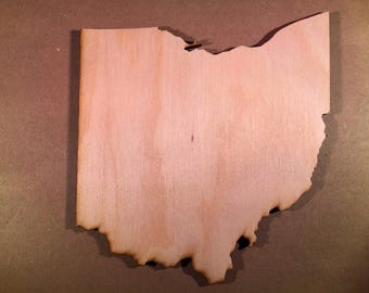 May include: A flat, light-colored wooden cutout shaped like the state of Ohio. The piece features a smooth, natural wood grain surface with darkened, laser-cut edges that define the irregular border of the state's geographic outline.