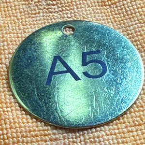 May include: A circular, metallic tag with a hole at the top. The tag has a brushed finish and features the bold text "A5" in a dark blue colour. The tag is slightly reflective.