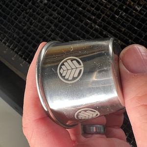 May include: A silver napkin ring with a leaf design etched on the surface. The ring is held in a hand, and the design is repeated on the ring. The napkin ring is made of metal.