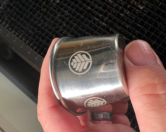 May include: A silver napkin ring with a leaf design etched on the surface. The ring is held in a hand, and the design is repeated on the ring. The napkin ring is made of metal.