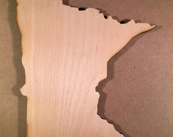 May include: A flat, light-colored wooden cutout shaped like the state of Minnesota. The piece features a smooth, natural wood grain surface with darkened, laser-cut edges that define the geographic outline of the state's borders.