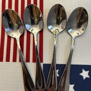 May include: A set of four polished silver-toned metal spoons arranged side by side. Each spoon features a smooth, rounded bowl and a plain, tapered handle. The reflective surface of the metal shows light highlights and shadows, creating a clean look.