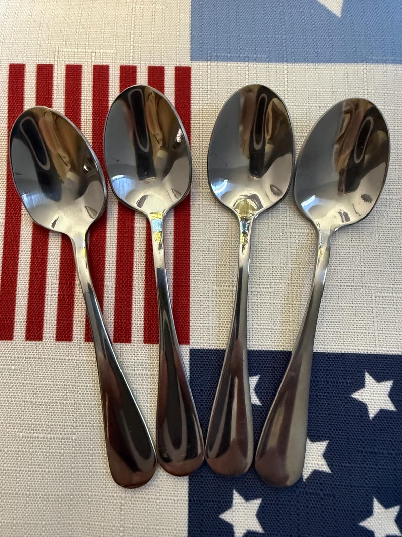 May include: A set of four polished silver-toned metal spoons arranged side by side. Each spoon features a smooth, rounded bowl and a plain, tapered handle. The reflective surface of the metal shows light highlights and shadows, creating a clean look.