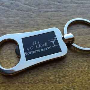 May include: A silver-colored bottle opener keychain with a black rectangular insert. The insert features the phrase "It's 5 O'Clock Somewhere!" and a martini glass graphic. The keychain is on a wooden surface.
