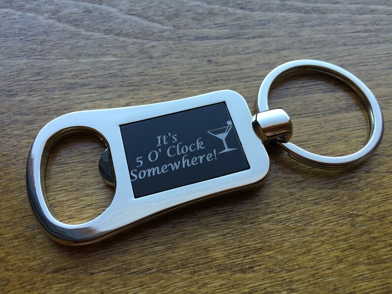 May include: A silver-colored bottle opener keychain with a black rectangular insert. The insert features the phrase "It's 5 O'Clock Somewhere!" and a martini glass graphic. The keychain is on a wooden surface.