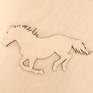 May include: A flat, light-coloured wooden cutout in the shape of a galloping horse. The silhouette features a detailed mane and tail with smooth, laser-cut edges, showcasing the natural grain and texture of the light beige wood material.