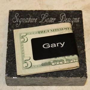 Pu&ograve; includere: Un fermasoldi nero con il nome "Gary" in lettere bianche, che tiene una banconota da cinque dollari. La clip poggia su un blocco di pietra grigio scuro con il testo "Signature Laser Designs" inciso.
