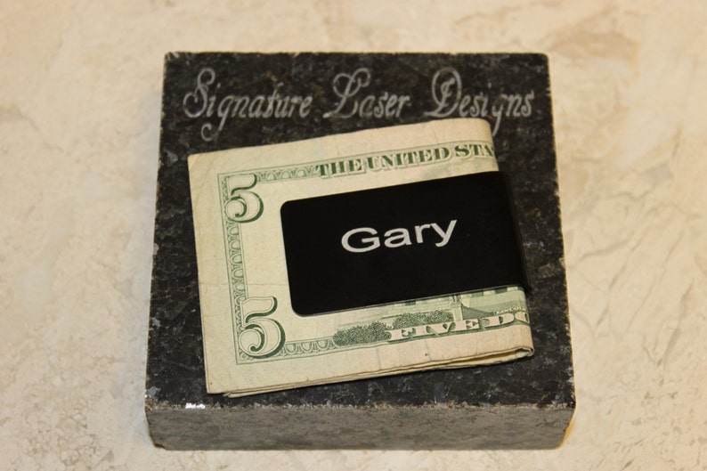 Pu&ograve; includere: Un fermasoldi nero con il nome "Gary" in lettere bianche, che tiene una banconota da cinque dollari. La clip poggia su un blocco di pietra grigio scuro con il testo "Signature Laser Designs" inciso.