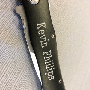 Może przedstawiać: Czarny scyzoryk ze srebrnym ostrzem i akcentami. Na rękojeści noża wygrawerowano imię "Kevin Phillips" w kolorze srebrnym. N&oacute;ż jest zamknięty i spoczywa na szarej powierzchni.