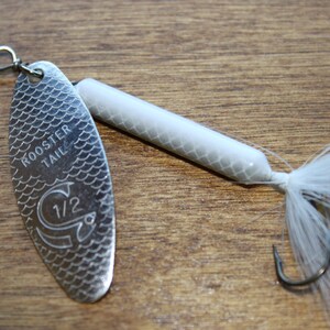 Op de afbeelding: Een zilveren Rooster Tail vislokmiddel met een witte body en staartveren. Het lokmiddel heeft een schubbenpatroon op het blad en de tekst "ROOSTER TAIL" en "1/2" zijn zichtbaar.