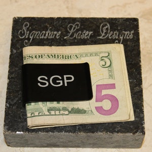 Pu&ograve; includere: Un fermasoldi nero con le iniziali "SGP" tiene una banconota da cinque dollari. La clip poggia su un blocco di pietra grigio scuro con il testo inciso "Signature Laser Designs". Lo sfondo &egrave; beige chiaro.