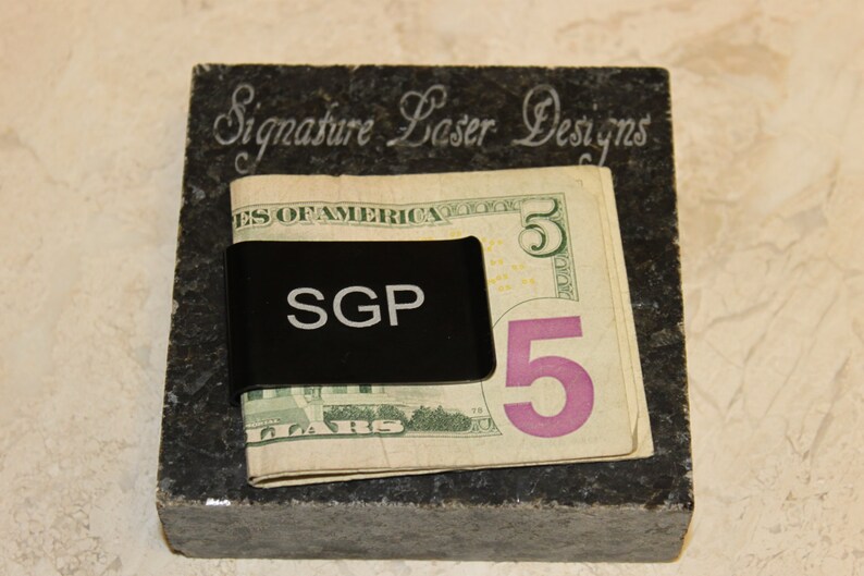 Pu&ograve; includere: Un fermasoldi nero con le iniziali "SGP" tiene una banconota da cinque dollari. La clip poggia su un blocco di pietra grigio scuro con il testo inciso "Signature Laser Designs". Lo sfondo &egrave; beige chiaro.