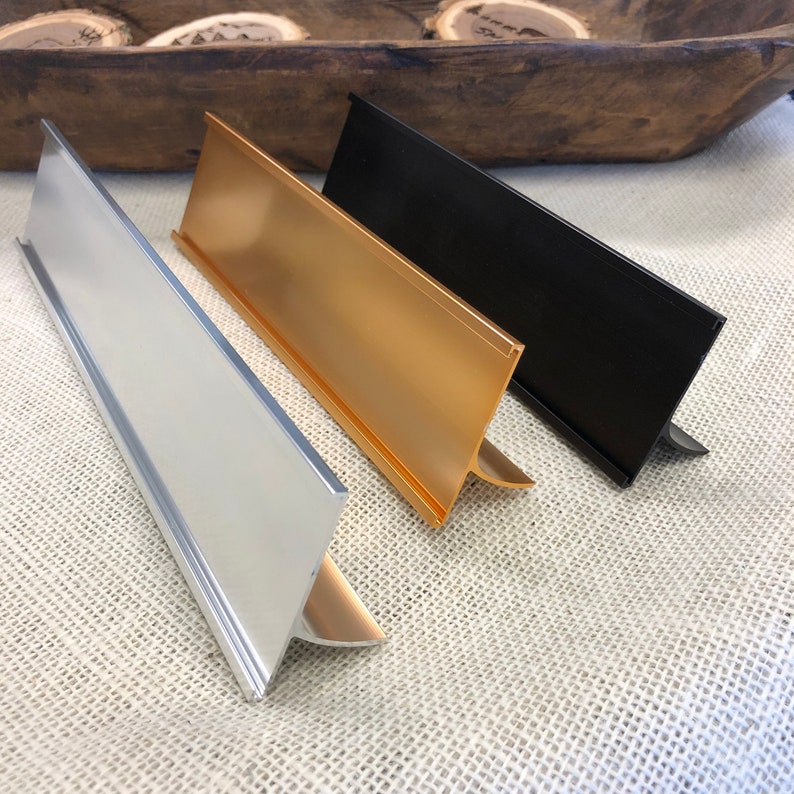 May include: Three metal desk nameplates in silver, gold, and black. Each nameplate has a triangular shape with a slot for a name card. The nameplates are displayed on a textured, neutral-coloured surface.