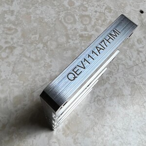 May include: A silver-colored metal component with the engraved text "QEV111AI7HMI". The component has a rectangular shape with a series of parallel fins on one side. The item is likely a heat sink or similar electronic part.