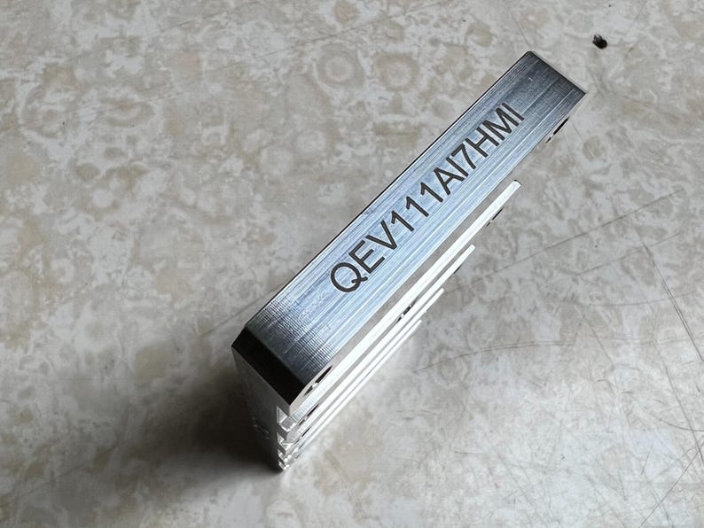 May include: A silver-colored metal component with the engraved text "QEV111AI7HMI". The component has a rectangular shape with a series of parallel fins on one side. The item is likely a heat sink or similar electronic part.