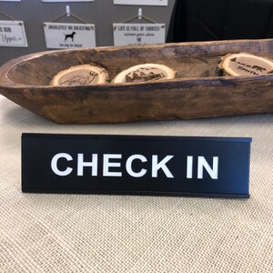 May include: A black rectangular sign with the words "CHECK IN" in white block letters. Behind the sign is a wooden bowl holding three small, round wooden slices with engraved designs. The bowl is dark brown.
