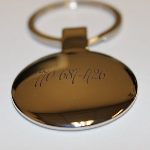May include: A shiny, metallic oval keychain with a silver ring. The keychain has the engraved phone number 770-687-1126. The background is a plain white surface.
