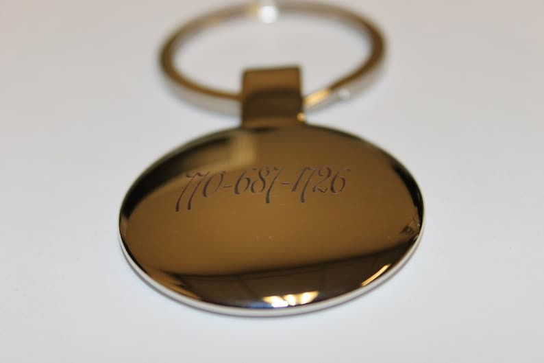 May include: A shiny, metallic oval keychain with a silver ring. The keychain has the engraved phone number 770-687-1126. The background is a plain white surface.