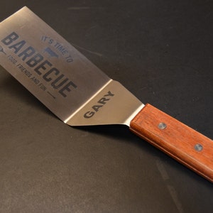 May include: A stainless steel spatula with a wooden handle secured by three rivets. The metal blade features the engraved text IT'S TIME TO BARBECUE, FOOD, FRIENDS AND FUN, and the name GARY, along with a small graphic of a spatula and a steak.