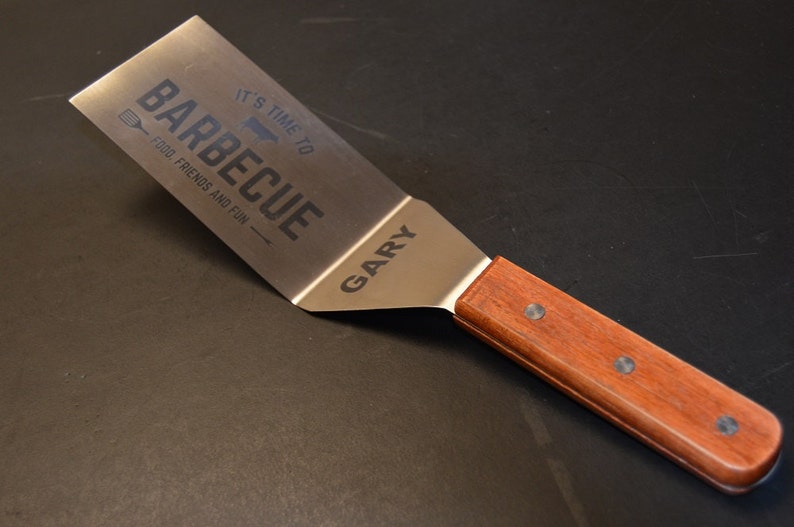 May include: A stainless steel spatula with a wooden handle secured by three rivets. The metal blade features the engraved text IT'S TIME TO BARBECUE, FOOD, FRIENDS AND FUN, and the name GARY, along with a small graphic of a spatula and a steak.