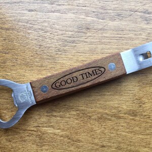 May include: A bottle opener with a wooden handle and stainless steel opener. The handle is engraved with the words "GOOD TIMES" in a black oval. The opener is made of stainless steel.