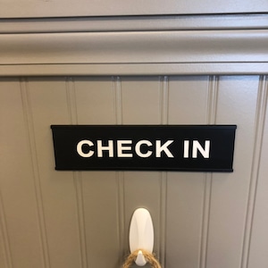 May include: A black sign with the words "CHECK IN" in white lettering is mounted on a gray wall. Below the sign is a white hook with a rope hanging from it. The wall has vertical paneling.