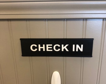 May include: A black sign with the words "CHECK IN" in white lettering is mounted on a gray wall. Below the sign is a white hook with a rope hanging from it. The wall has vertical paneling.