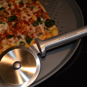 Op de afbeelding: Een rechthoekige pizza met beleg, waaronder broccoli, wordt gesneden met een roestvrijstalen pizzasnijder. De pizzasnijder heeft de naam "Anita" op de handgreep gegraveerd. De pizza ligt op een ronde, grijze pizzapan.