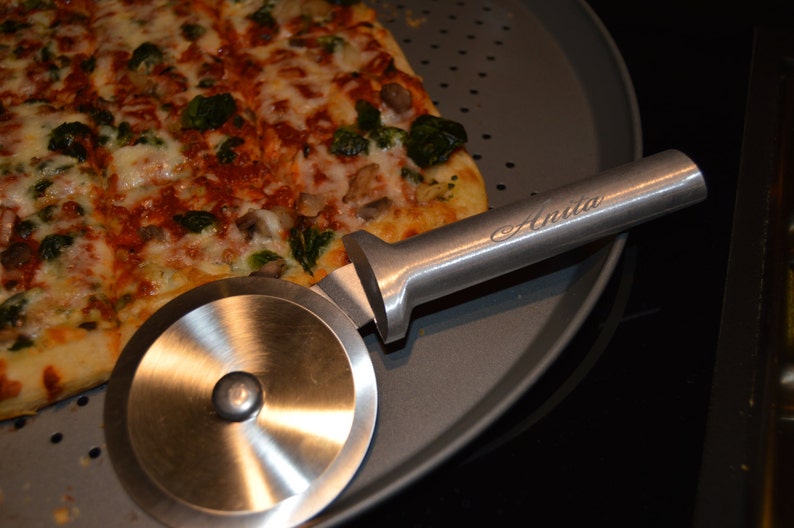 Op de afbeelding: Een rechthoekige pizza met beleg, waaronder broccoli, wordt gesneden met een roestvrijstalen pizzasnijder. De pizzasnijder heeft de naam "Anita" op de handgreep gegraveerd. De pizza ligt op een ronde, grijze pizzapan.