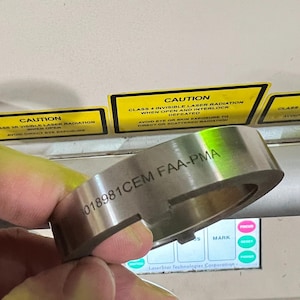 Op de afbeelding: Een zilveren metalen ring met de tekst "018981CEM FAA-PMA" gegraveerd op het oppervlak. De ring wordt in een hand gehouden, met waarschuwingslabels zichtbaar op de achtergrond. De labels waarschuwen voor laserstraling.
